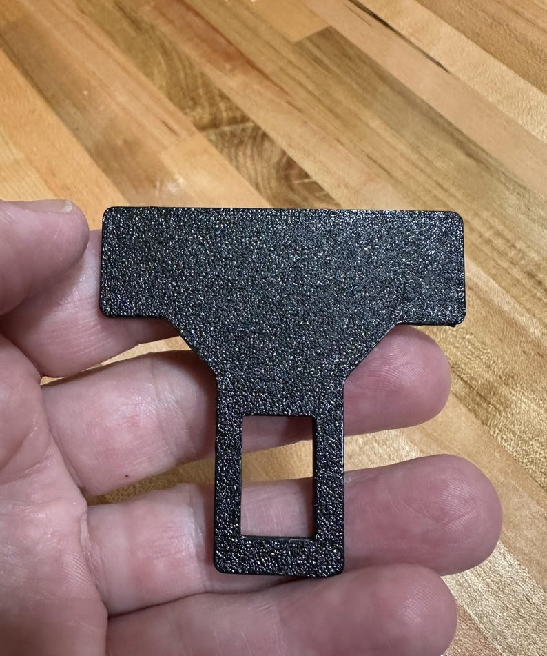 May include: A black seat belt buckle insert with a textured, matte finish. The object features a wide, rectangular top section that tapers down into a narrower, vertical stem containing a rectangular cutout opening in the center of the lower portion.
