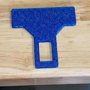 May include: A blue seat belt buckle insert with a glittery finish. The object features a wide rectangular top section that tapers down into a narrower base with a central rectangular cutout, designed to fit into a standard vehicle seat belt receiver.