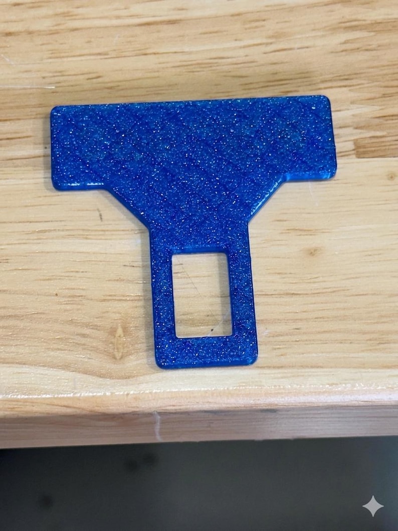 May include: A blue seat belt buckle insert with a glittery finish. The object features a wide rectangular top section that tapers down into a narrower base with a central rectangular cutout, designed to fit into a standard vehicle seat belt receiver.