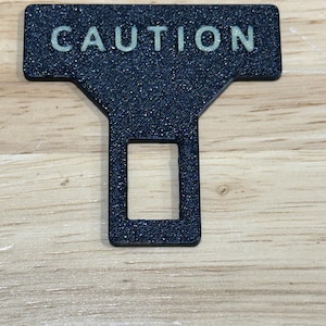 May include: A black T-shaped seat belt buckle insert with a textured finish and the word CAUTION printed in light-colored capital letters across the top horizontal bar, featuring a rectangular cutout in the lower vertical section.