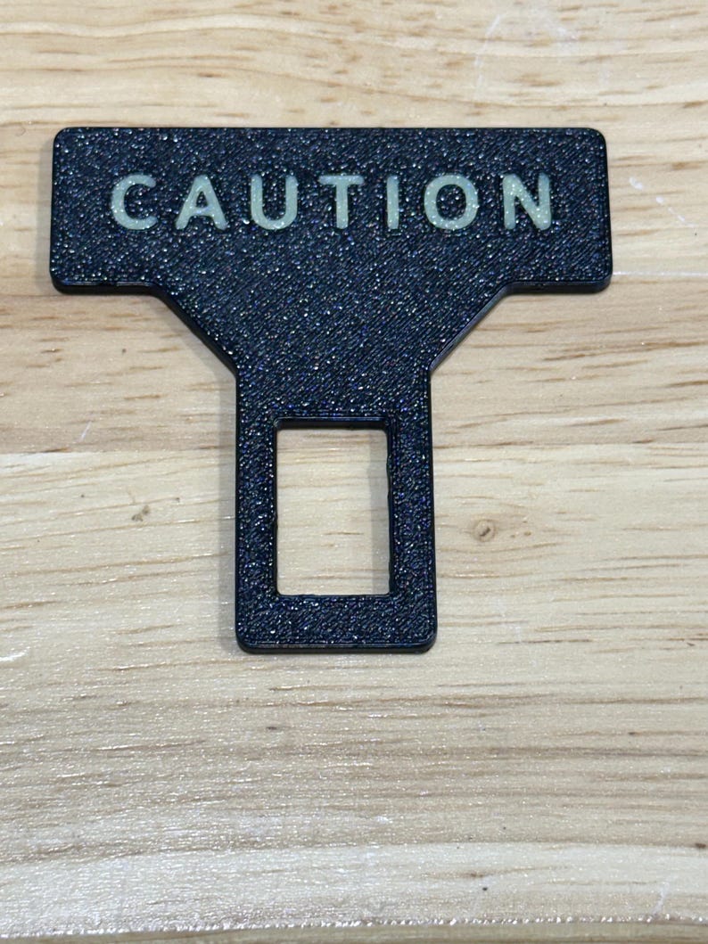 May include: A black T-shaped seat belt buckle insert with a textured finish and the word CAUTION printed in light-colored capital letters across the top horizontal bar, featuring a rectangular cutout in the lower vertical section.