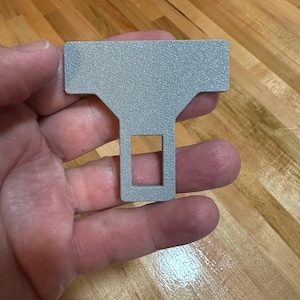 May include: A silver-colored metal seat belt buckle extender with a textured, matte finish. The object features a wide, rectangular top section that tapers down into a narrower, vertical stem containing a rectangular cutout in the center.