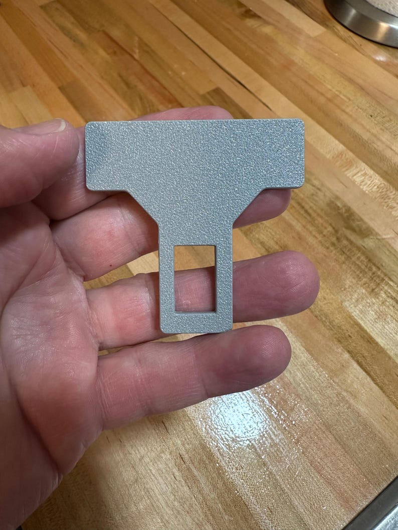 May include: A silver-colored metal seat belt buckle extender with a textured, matte finish. The object features a wide, rectangular top section that tapers down into a narrower, vertical stem containing a rectangular cutout in the center.