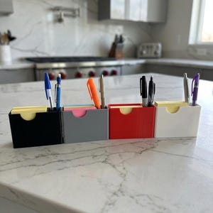 Puede incluir: Un conjunto de cuatro organizadores de escritorio rectangulares en negro, gris, rojo y blanco. Cada organizador cuenta con una abertura frontal para acceder fácilmente a notas adhesivas y una ranura dedicada para bolígrafos o lápices.