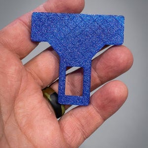 May include: A T-shaped seat belt buckle silencer made of blue glittery material. The object features a textured, diamond-patterned surface on the top horizontal bar and a rectangular cutout in the lower vertical section, designed for insertion.