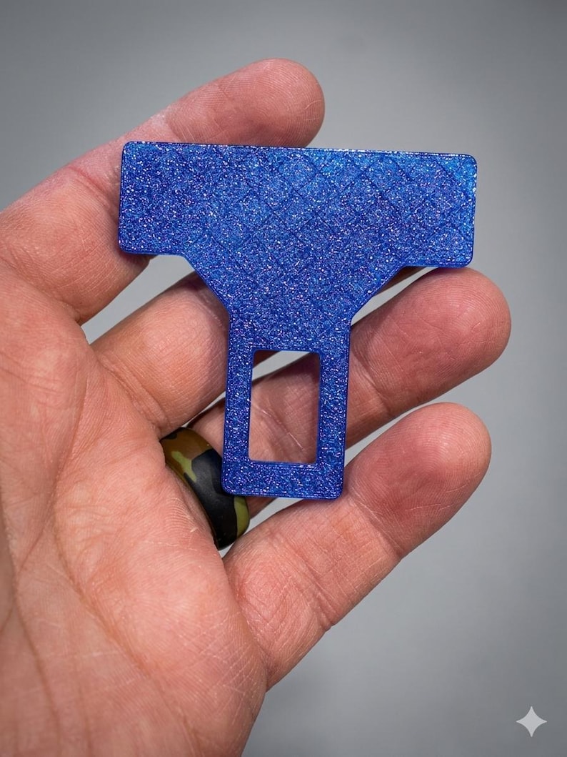 May include: A T-shaped seat belt buckle silencer made of blue glittery material. The object features a textured, diamond-patterned surface on the top horizontal bar and a rectangular cutout in the lower vertical section, designed for insertion.