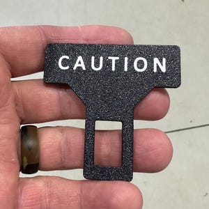 May include: A black T-shaped seat belt buckle insert with a textured, granular surface finish. The word CAUTION is printed in white, capitalized, sans-serif letters across the top horizontal bar. A rectangular opening is centered in the lower section.
