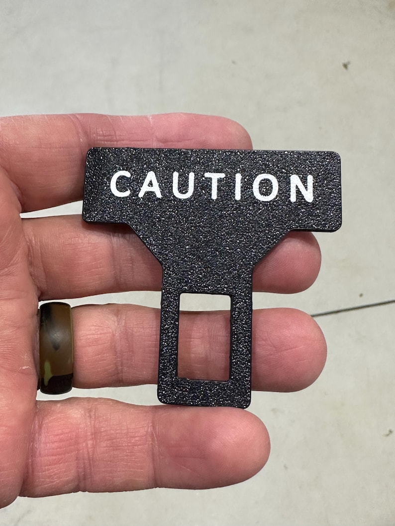 May include: A black T-shaped seat belt buckle insert with a textured, granular surface finish. The word CAUTION is printed in white, capitalized, sans-serif letters across the top horizontal bar. A rectangular opening is centered in the lower section.