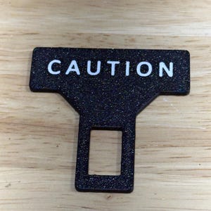 May include: A black, glittery seat belt buckle with the word "CAUTION" in white capital letters. The buckle has a rectangular opening and a T-shaped top. The background is a light brown wooden surface.