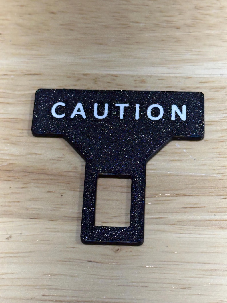 May include: A black T-shaped seat belt buckle extender clip with a textured surface and the word CAUTION printed in white capital letters across the top horizontal bar. The clip features a rectangular cutout in the center of the vertical stem.