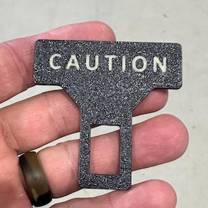 May include: A black seat belt buckle insert with a textured, glittery finish. The T-shaped object features the word CAUTION printed in white capital letters across the top horizontal bar and a rectangular cutout in the lower vertical section.