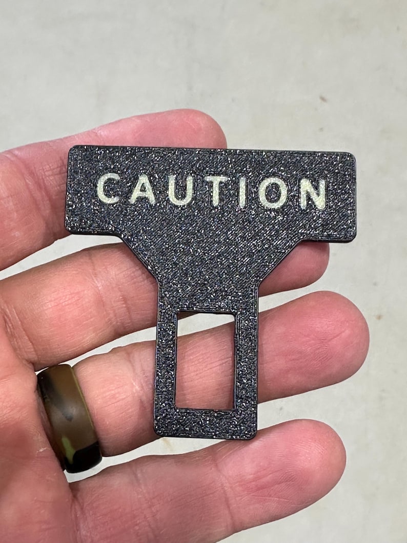 May include: A black seat belt buckle insert with a textured, glittery finish. The T-shaped object features the word CAUTION printed in white capital letters across the top horizontal bar and a rectangular cutout in the lower vertical section.