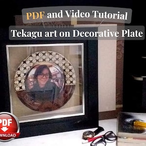 Op de afbeelding: Een ingelijste decoratieve plaat met Tekagu-kunst. De plaat heeft een bruin en wit patroon en een centraal beeld van een persoon. Het zwarte frame omringt de plaat. De afbeelding bevat de tekst "PDF and Video Tutorial Tekagu art on Decorative Plate."