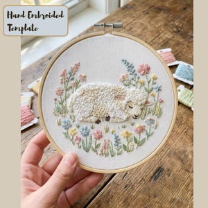 May include: Hand-embroidered template featuring a sleeping sheep surrounded by colorful flowers. The artwork is set in a wooden embroidery hoop. The text "Hand Embroidered Template" is in the upper left corner. The color palette includes cream, beige, pink, blue, and green.