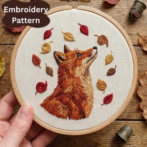 May include: An embroidery pattern showing a fox with orange and brown fur, gazing upwards. The design includes embroidered autumn leaves in red, gold, and brown, set against cream fabric within a wooden hoop. The text "Embroidery Pattern" is in a brown oval.