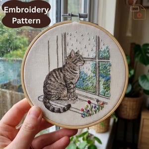 May include: An embroidery hoop with a tabby cat design looking out of a window at the rain. The text "Embroidery Pattern" is in a brown oval. The cat sits on a windowsill with embroidered flowers below. The hoop is held by a hand.