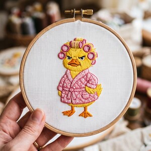 Pode incluir: Um bastidor de bordado circular de madeira contendo um painel de tecido branco com um desenho bordado à mão de um patinho amarelo. O patinho usa bobes cor-de-rosa e um roupão acolchoado rosa com bolinhas brancas, com expressão rabugenta.