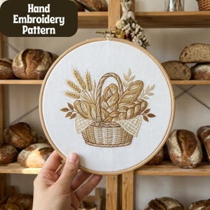 Op de afbeelding: Handborduurpatroon met een mand vol brood en tarwearen. De borduursels zijn in bruine en beige tinten op een witte achtergrond, in een houten ring. De tekst "Hand Embroidery Pattern" staat linksboven.