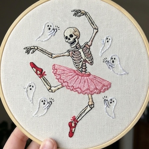 May include: An embroidered hoop art piece featuring a skeleton ballerina in a pink tutu and red ballet shoes. The skeleton is surrounded by white ghost figures. The artwork is set against a neutral fabric background and framed by a wooden hoop.