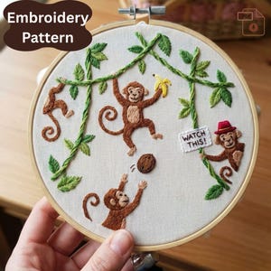 May include: Embroidery pattern featuring a playful monkey scene. Brown monkeys are embroidered on a cream-colored fabric, with green vines and leaves. One monkey holds a banana, another a sign that says "WATCH THIS!" The design is set in a wooden hoop.
