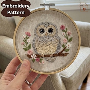 Peut inclure: Broderie d'un hibou dans un cercle en bois. Le hibou, aux tons gris et beige, est perché sur une branche marron, entouré de fleurs roses et blanches. Le texte "Embroidery Pattern" est visible en haut à gauche.