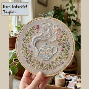 May include: Hand-embroidered template featuring a teapot design. The teapot is light blue with floral patterns, surrounded by a wreath of colorful flowers and greenery. The text "Hand Embroidered Template" is at the top. The embroidery is set in a wooden hoop.