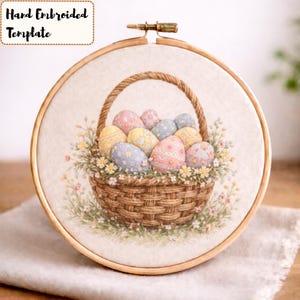 May include: Hand-embroidered Easter egg basket template in a wooden hoop. The basket is filled with pastel-colored eggs and surrounded by flowers. The text "Hand Embroidered Template" is in a cream-colored box.