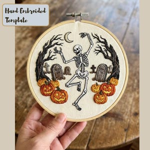 Puede incluir: Arte de Halloween bordado a mano en un aro, que representa un esqueleto bailando, calabazas y lápidas. El diseño incluye una luna creciente y árboles desnudos, todo dentro de un aro de bordado de madera. El texto "Hand Embroidered Template" está en la esquina superior izquierda.