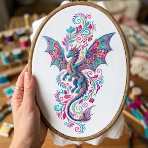 May include: An oval wooden embroidery hoop containing white fabric with a detailed, colorful thread-stitched dragon. The dragon features vibrant blue, pink, and gold patterns, surrounded by intricate floral and swirling decorative embroidery.