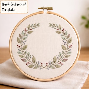 May include: A round embroidery hoop with a cream-colored fabric featuring a hand-embroidered wreath design. The wreath includes green leaves, red and blue berries, and the text "Hand Embroidered Template" in a cream-colored box.