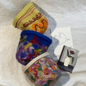 Peut inclure: Trois paniers en laine feutrée aux motifs jaune, bleu et multicolore. Le panier jaune présente des détails brodés. Une carte avec une illustration de colombe et une peinture d'un vase avec une fleur sont également présentes.