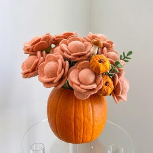 Peut inclure: Un bouquet de fleurs en feutrine couleur pêche disposé dans une citrouille orange vif. L'arrangement comprend deux petites citrouilles en feutrine orange, un champignon en feutrine avec un chapeau marron et de petites feuilles vertes.