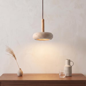 Könnte beinhalten: Eine moderne Pendelleuchte mit beigem, gesprenkeltem Schirm und Holzakzent. Das Licht hängt an einem braunen Kabel. Auf einem Holztisch stehen eine Vase mit getrocknetem Gras, ein Krug und ein Glas Wasser. Der Hintergrund ist eine strukturierte weiße Wand.