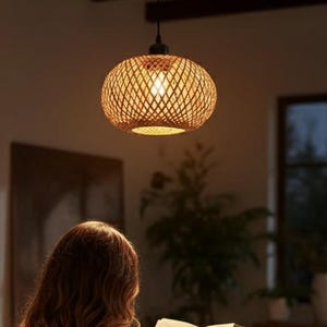 Puede incluir: Una escena interior cálida y acogedora con una lámpara colgante de ratán tejido sobre una persona leyendo un libro en un sillón. Una estantería de madera y una mesita auxiliar con una taza contribuyen al ambiente confortable. La luz emite un brillo suave.