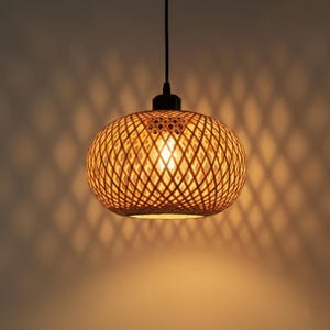 Peut inclure: Un luminaire suspendu sphérique avec un abat-jour en bambou tressé brun clair. La lumière est allumée, dégageant une lueur chaude. Le luminaire est suspendu à un cordon et un auvent noirs.