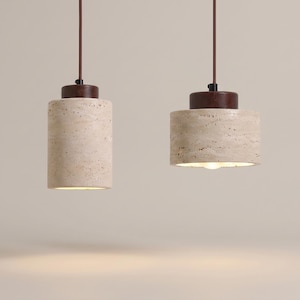 Może przedstawiać: Dwie lampy wiszące z cylindrycznymi kloszami wykonanymi z beżowego, porowatego kamienia trawertynowego. Każda lampa posiada ciemnobrązową drewnianą nasadkę u góry i jest zawieszona na cienkim brązowym przewodzie. Emitują ciepłe światło od dołu.