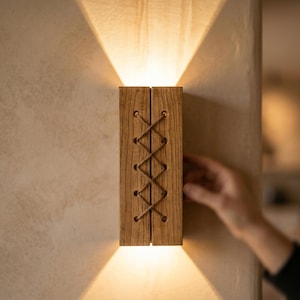 May include: Wooden wall sconce emitting a warm light. The sconce is constructed from two wooden panels, joined with a decorative crisscross pattern. The light shines upwards and downwards, creating a unique lighting effect. The sconce is mounted on a textured wall.