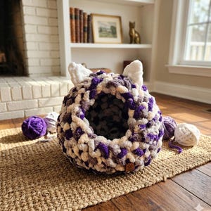 May include: A handmade cat bed in a round shape, featuring a mix of purple, white, and gray yarn. The bed has two white, ear-shaped accents on top. Several balls of yarn in matching colors are scattered around the bed, suggesting a cozy, handcrafted theme.