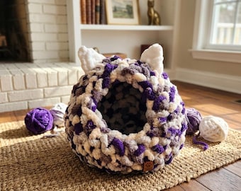 May include: A round, chunky crochet pet bed featuring a variegated pattern of purple, grey, and cream yarn. The bed has a circular opening and two small, white, triangular ears attached to the top. Several yarn balls in matching colors sit nearby.