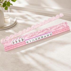 May include: A pink Mahjong tile rack with a scalloped edge and a pink marble design. The rack holds several Mahjong tiles with various symbols and numbers. The tiles are white with black and red markings. The rack is on a white surface.
