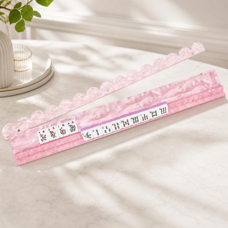 May include: A pink Mahjong tile rack with a scalloped edge and a pink marble design. The rack holds several Mahjong tiles with various symbols and numbers. The tiles are white with black and red markings. The rack is on a white surface.