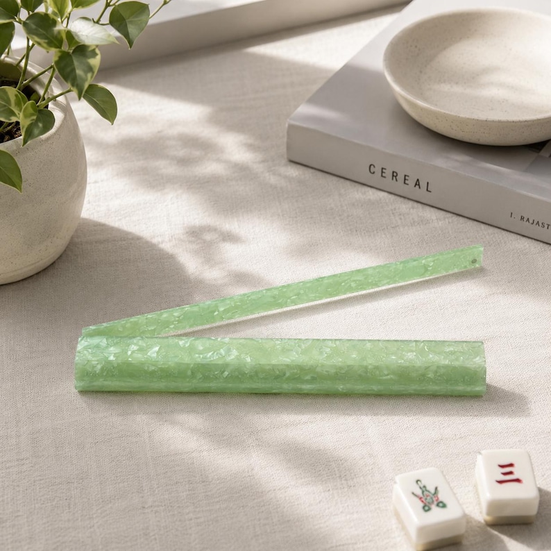 May include: A light green, segmented ruler made of a textured material, partially open on a light-colored surface. The word "CEREAL" is visible on a book in the background, along with a small bowl and a potted plant. Two Mahjong tiles are in the foreground.