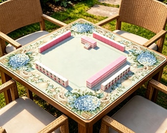 Puede incluir: Una mesa de mahjong cuadrada de madera con un diseño floral y cuatro sillas de mimbre a juego. La mesa presenta una superficie verde claro con detalles de hortensias azules y conchas marinas. Las fichas de mahjong rosas están dispuestas sobre la mesa.