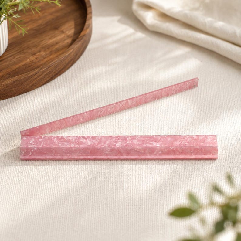May include: A pink, rectangular acrylic tray with a textured surface. The tray is open and angled, with one side raised. The tray is on a white surface, with a wooden tray and greenery in the background.