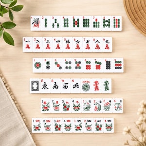 May include: A set of white Mahjong tiles with various symbols and numbers, including dragons, flowers, and dots. The tiles are arranged on a light wooden surface, with some greenery and a woven mat in the background.