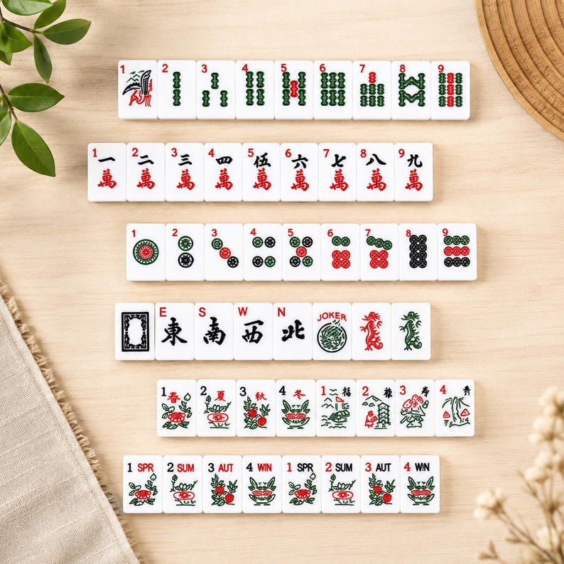 May include: A set of white Mahjong tiles with various symbols and numbers, including dragons, flowers, and dots. The tiles are arranged on a light wooden surface, with some greenery and a woven mat in the background.