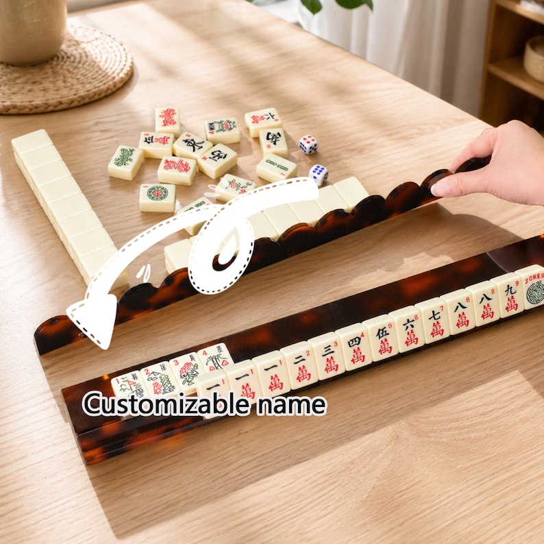 May include: A mahjong set with ivory-colored tiles and a brown, tortoise-shell patterned rack. The tiles have red and green symbols. The text "Customizable name" is visible on the rack. Dice and additional tiles are scattered on the wooden table.