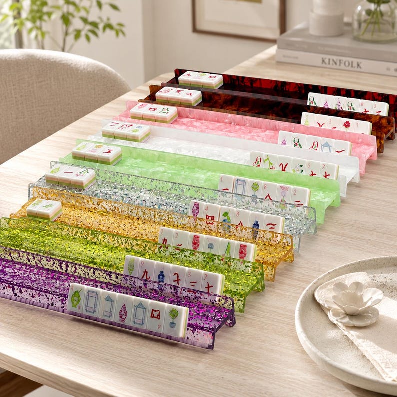 May include: A collection of colorful acrylic mahjong tile racks in various colors, including purple, gold, green, and pink. Each rack holds a set of mahjong tiles, showcasing the game's intricate designs. The racks are arranged on a light-colored wooden surface.
