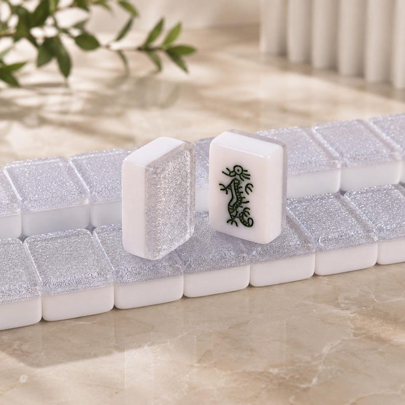 May include: Close-up of white and clear Mahjong tiles. One tile features a green dragon design. The tiles are arranged on a silver and white surface. The game pieces are rectangular and have a glossy finish.