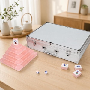 May include: A complete Mahjong set with pink tiles and a silver carrying case. The tiles are arranged in a pyramid shape, with dice and additional tiles scattered on the wooden surface. The set includes a variety of tiles with different symbols and characters.